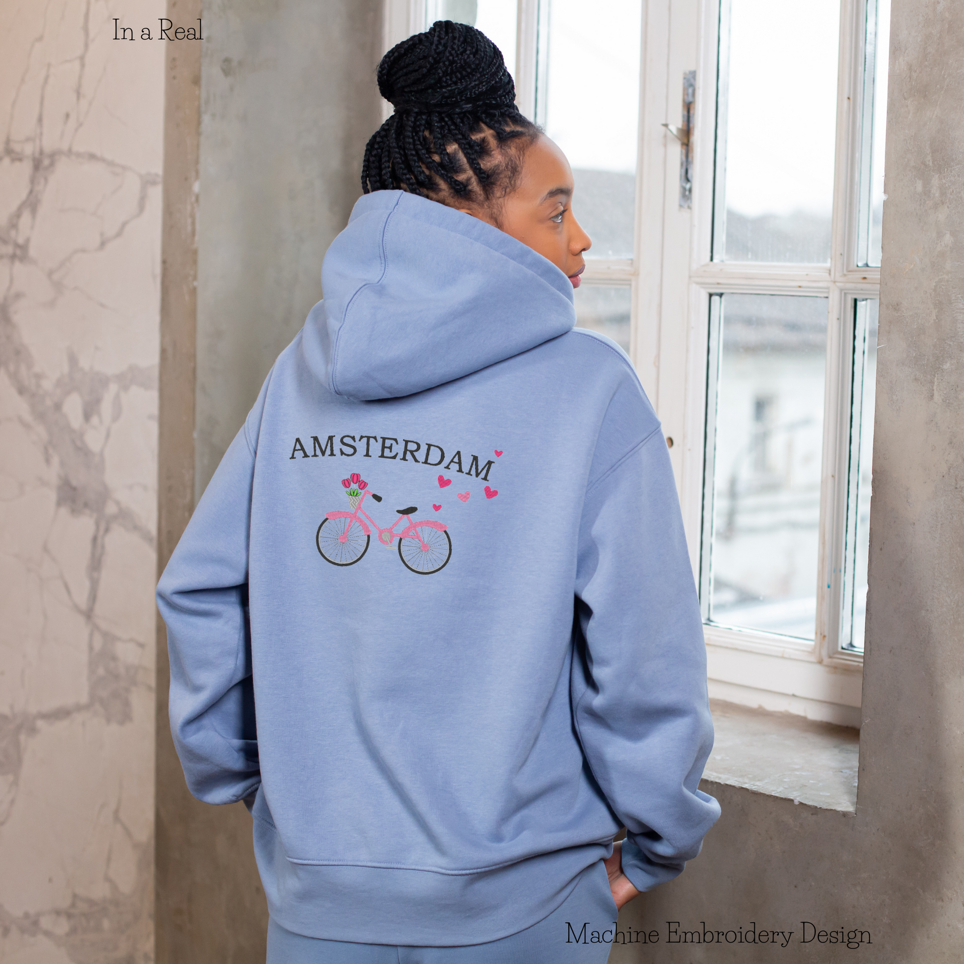 Machine embroidery design adorned with hearts, featuring the written phrase 'AMSTERDAM' and a bicycle carrying tulips in a basket, displayed on a hoodie