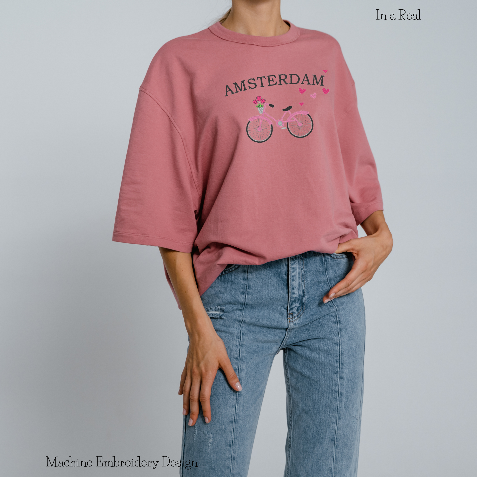 Machine embroidery design adorned with hearts, featuring the written phrase 'AMSTERDAM' and a bicycle carrying tulips in a basket, displayed on a T-shirt