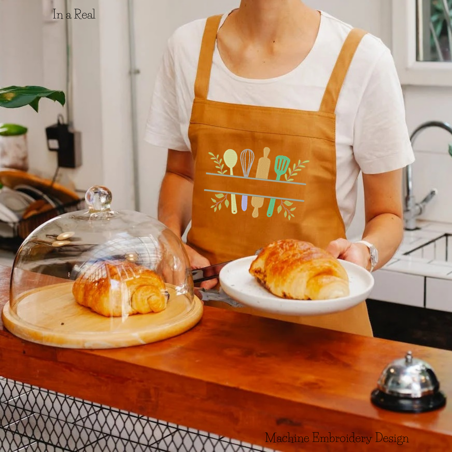 Intricately embroidered split-frame monogram surrounded by kitchen utensils, displayed on an apron