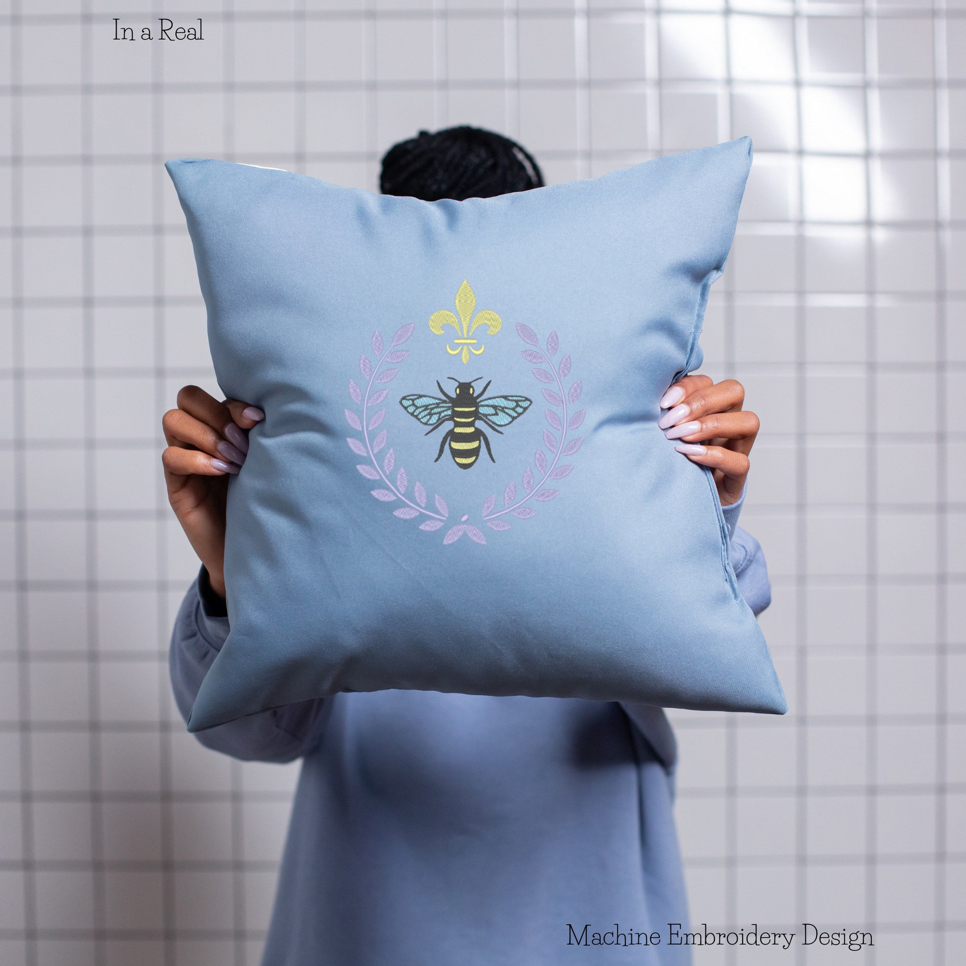 An intricate machine embroidery design featuring a regal bee surrounded by a laurel frame is displayed on a pillow