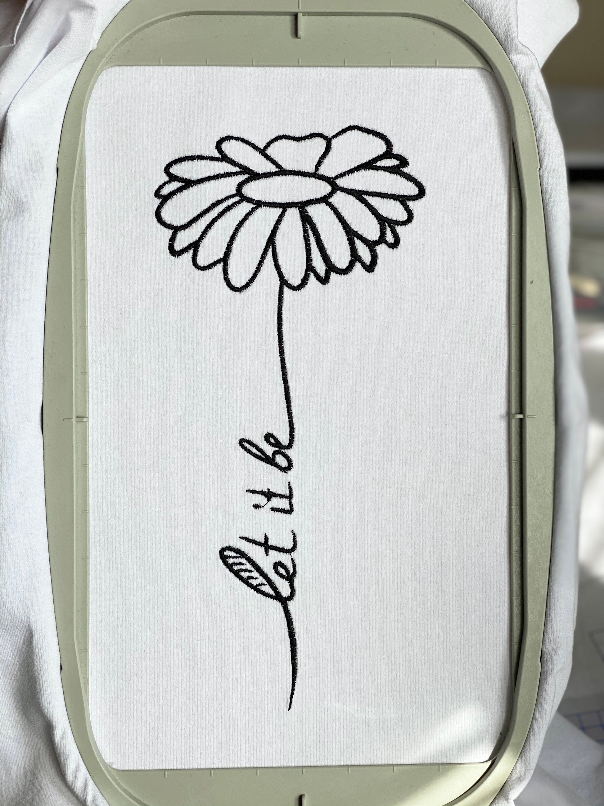 An image displaying an embroidery design where the words 'Let it be' are intertwined with a delicate chamomile flower